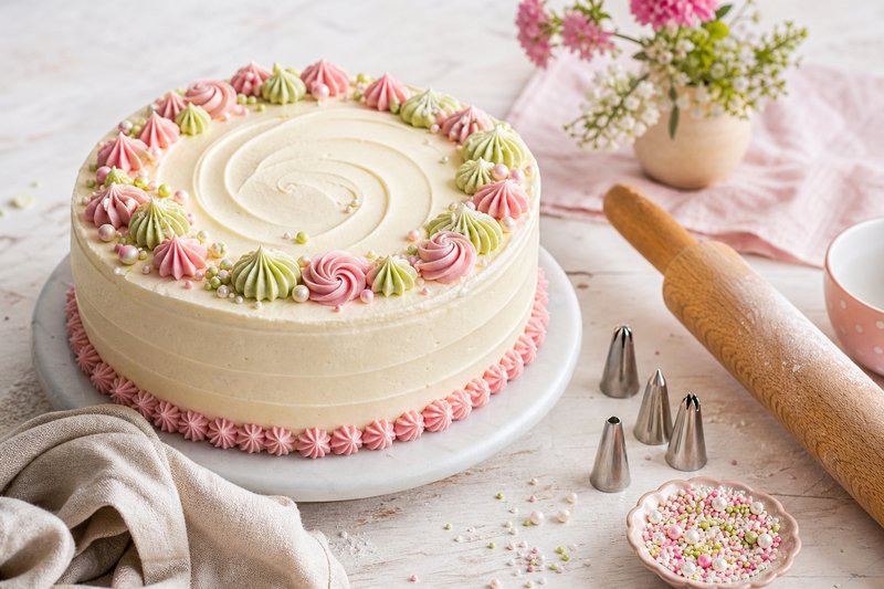 Frisch gefüllte Torte mit glatter Buttercreme, Spritztüllen und pastellfarbenen Zuckerperlen auf heller Arbeitsfläche